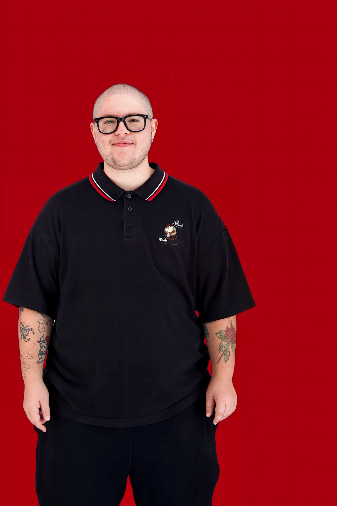 Smiling man with black glasses wearing a black polo shirt, tattoos visible, against a solid red background.